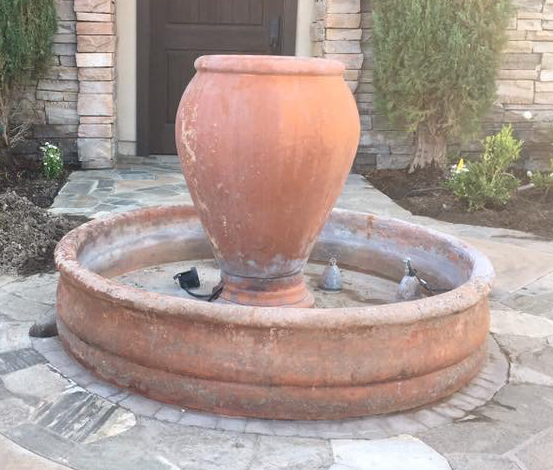 A large, terracotta-colored stone fountain with a central jar and circular basin, set on a stone patio outside a doorway.