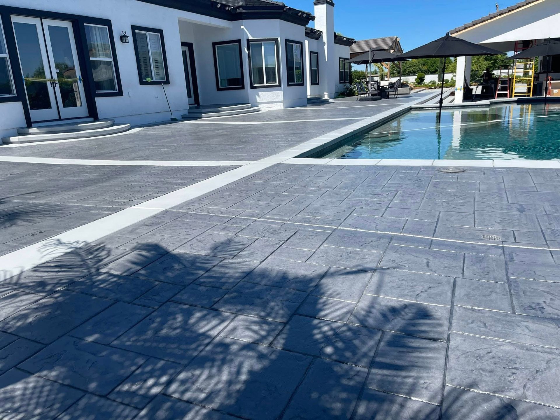 Exterior view of a patio with a pool; gray stamped concrete, white trim, and a house in the background.