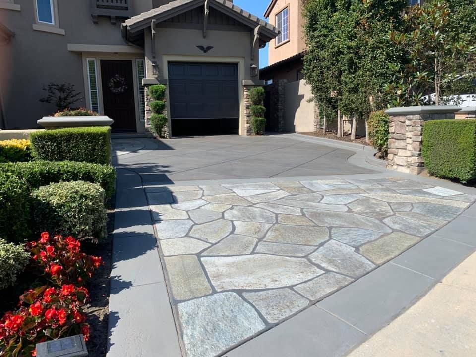 Stone driveway leading to a house with a gray garage door, bordered by green hedges and flowerbeds.