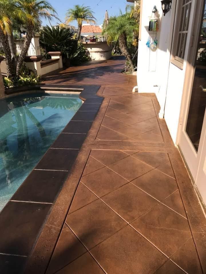Brown concrete patio with a pool, diagonal pattern, next to a white house with palm trees.
