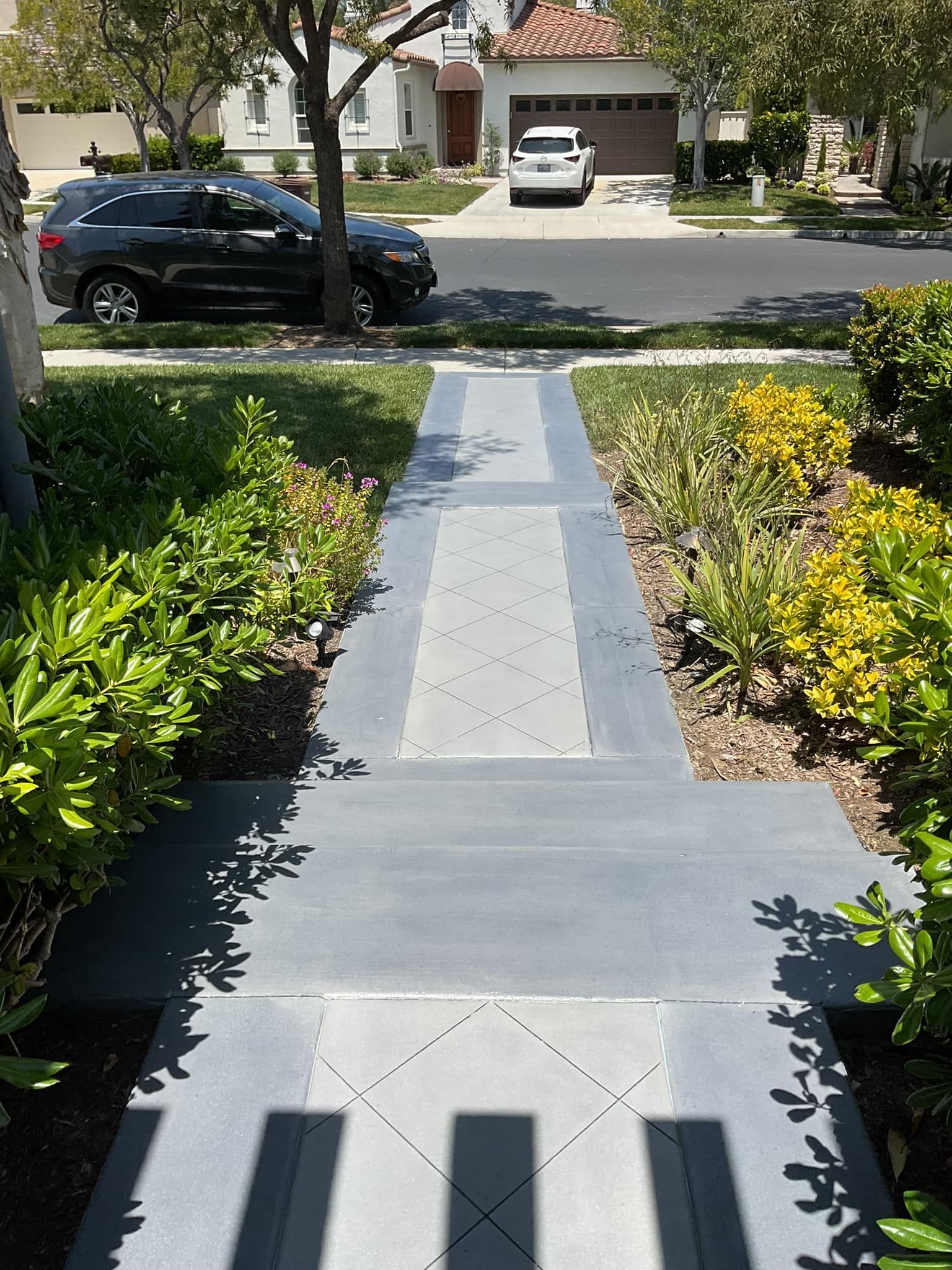 Concrete walkway with gray and blue accents leads to a house with parked cars on a sunny day.