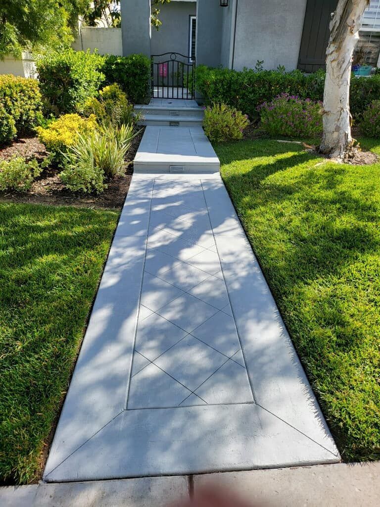Concrete walkway leading to a house with a black gate, lined with grass and greenery.