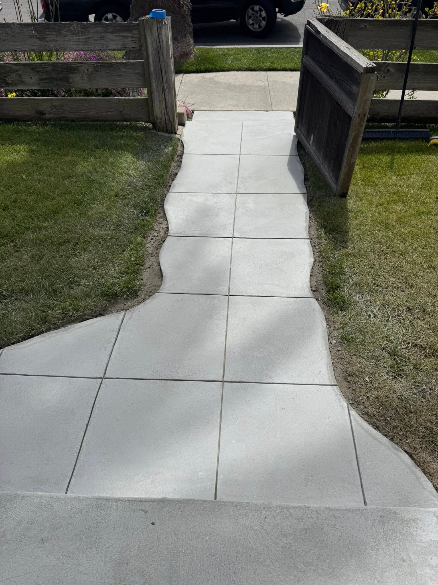 Concrete walkway leading from a yard to a house, bordered by grass and wooden fences.