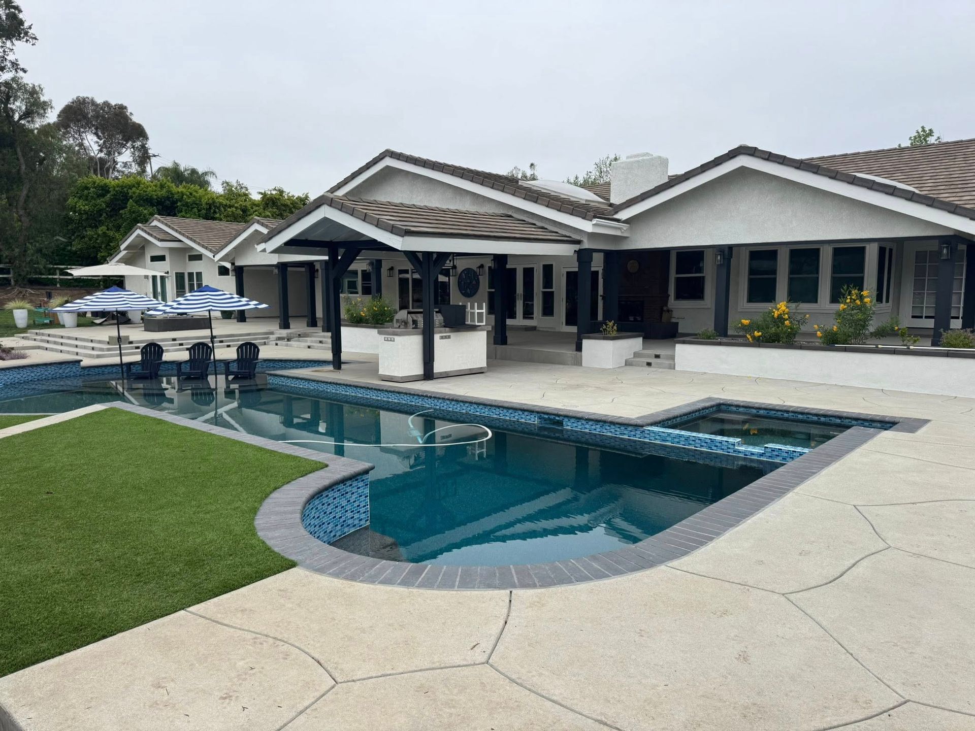 Pool and house exterior with an overcast sky; lush green lawn, chairs, and a patio.