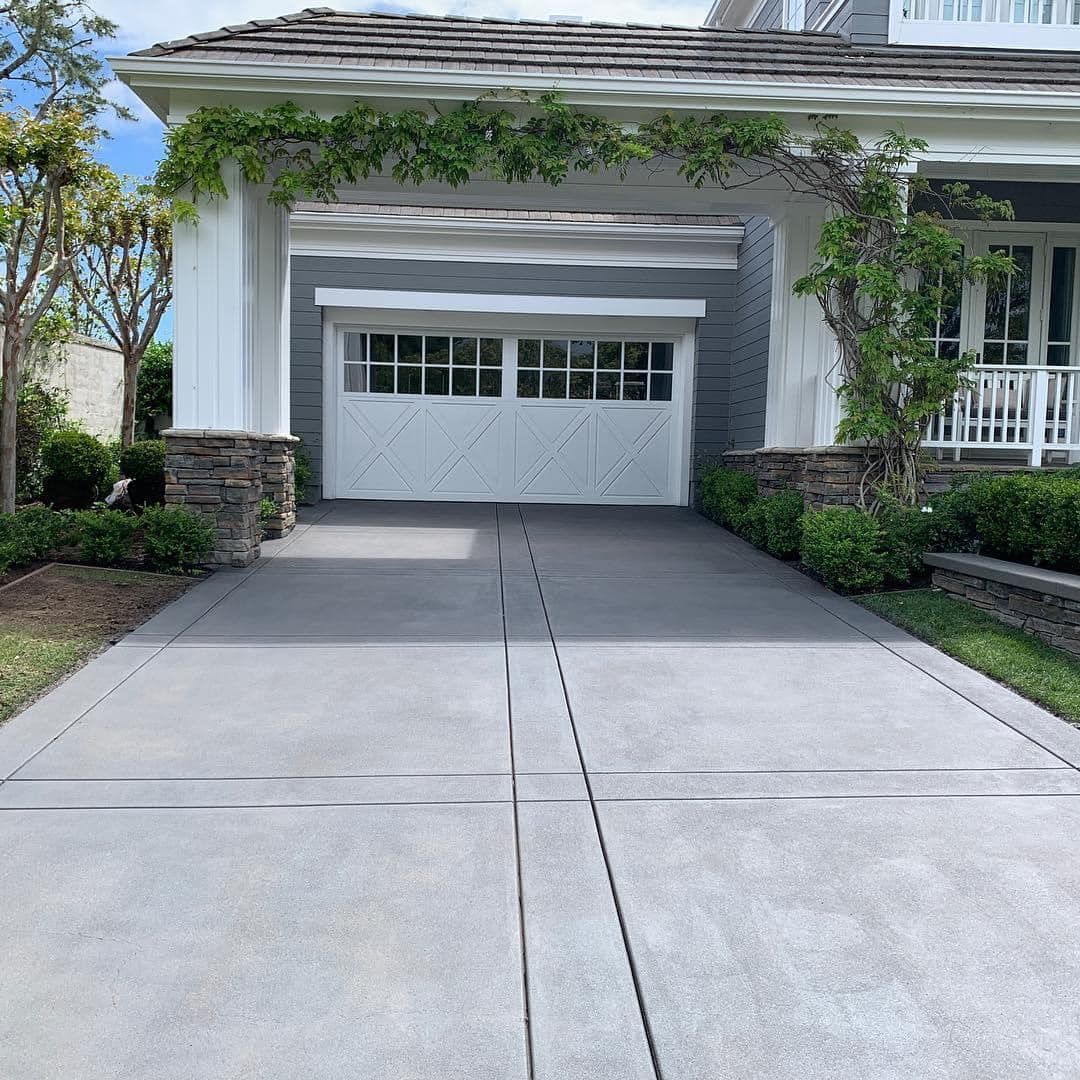 Driveway leading to a white garage with stone accents, lush greenery, and a house in the background.