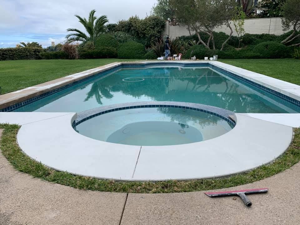 Swimming pool with a spa, surrounded by concrete and grass.