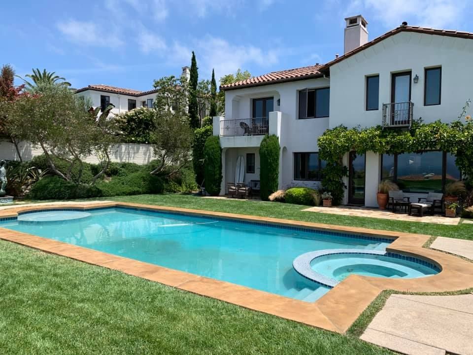 A large house with a pool and jacuzzi in a sunny backyard with green grass and a blue sky.