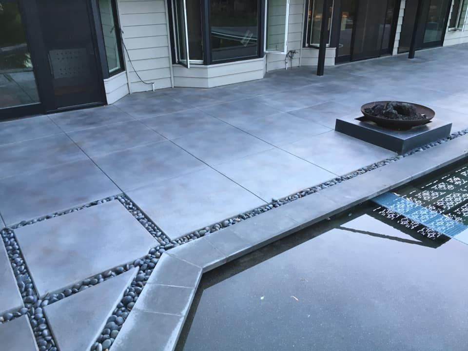 Gray concrete patio with black stone accents, adjacent to a pool and a contemporary home.