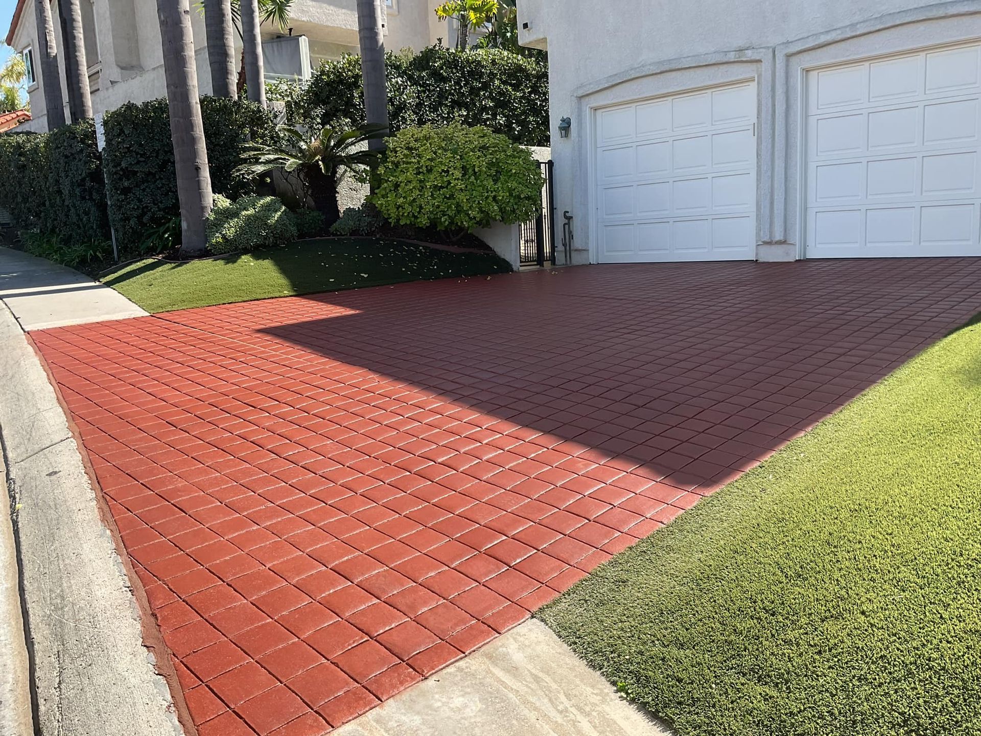 Red brick paved driveway in front of a white garage, next to green grass and a sidewalk.