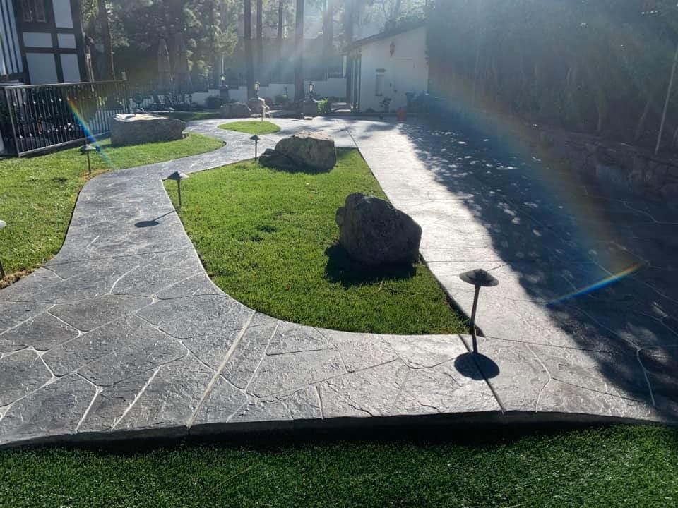 Pathway of gray textured concrete with green grass and two large rocks. Outdoor setting.