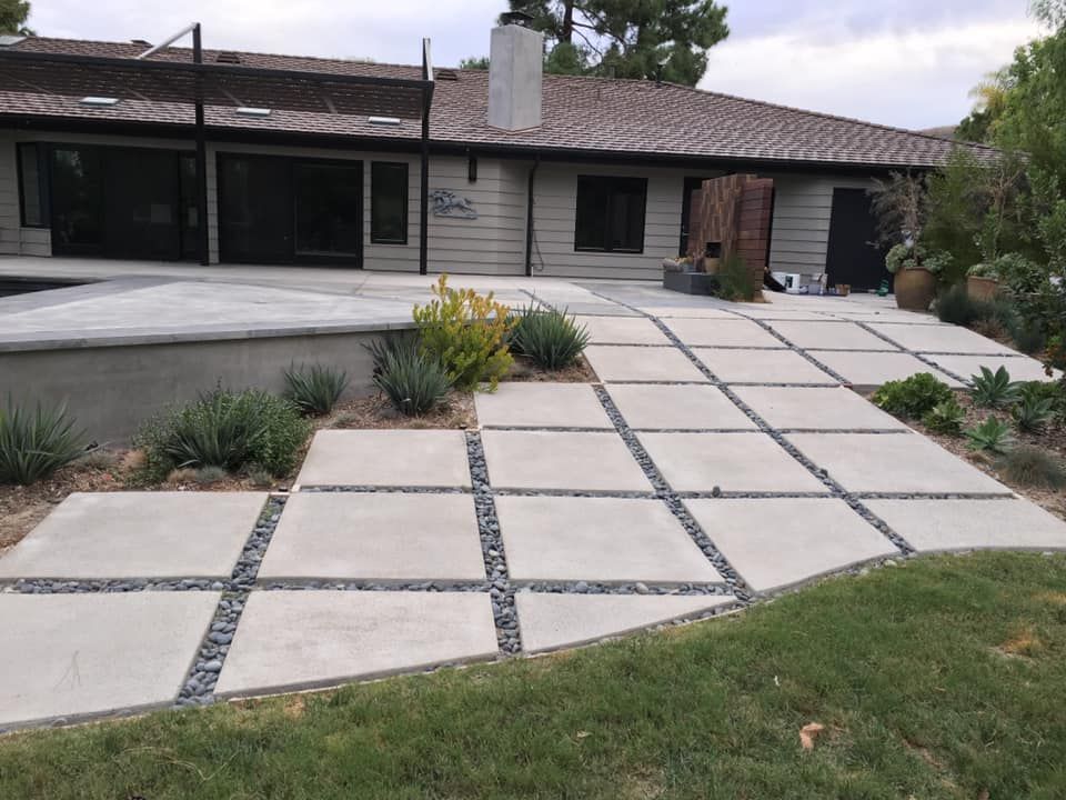 Concrete pavers with dark rock accents lead to a house with a pool deck, plants, and a grassy yard.