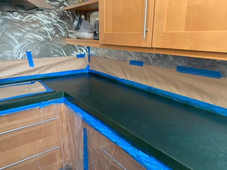 A dark green countertop in a kitchen, taped off and covered for painting, with light wood cabinets above.