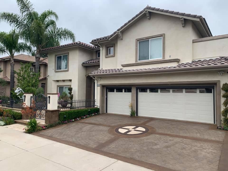 Beige two-story house with a two-car garage. Stamped concrete driveway with decorative design. Overcast sky.