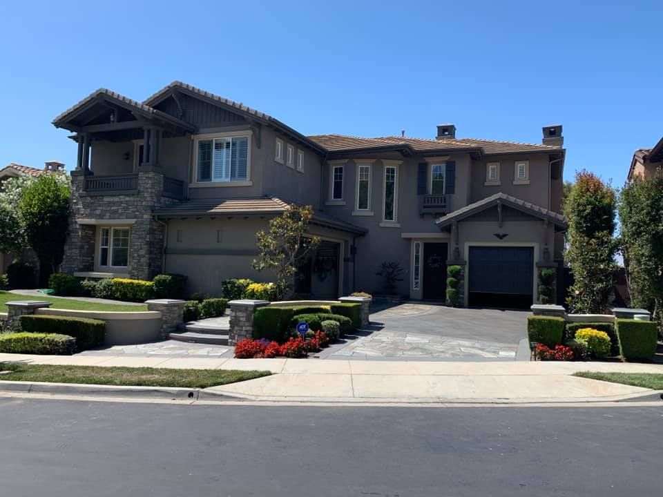 Two-story house with gray stucco, stone accents, and manicured landscaping, fronted by a paved driveway and street.