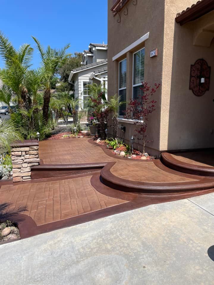 Brown stamped concrete walkway leading to a house with landscaping and palm trees.
