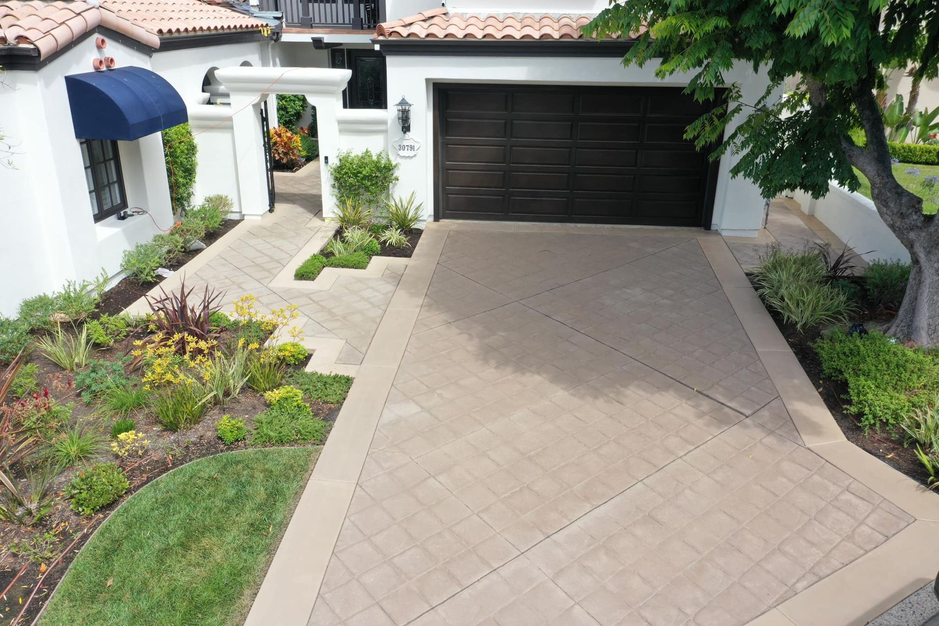 Driveway with brown garage door, landscaping, and pathway leading to white house with blue awning.