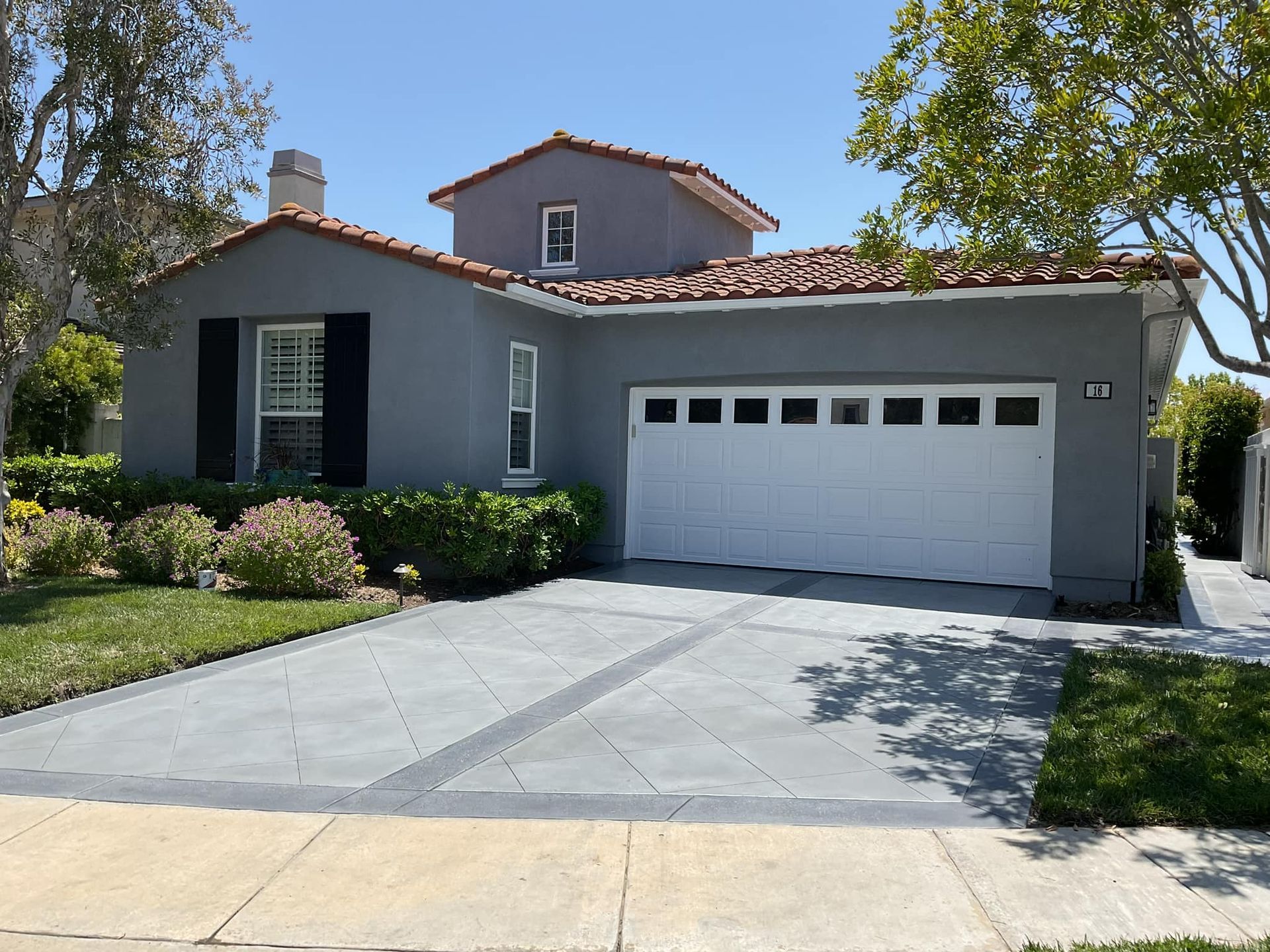 Gray house with white garage door, red-tiled roof, and gray driveway. Green lawn and trees surround it.
