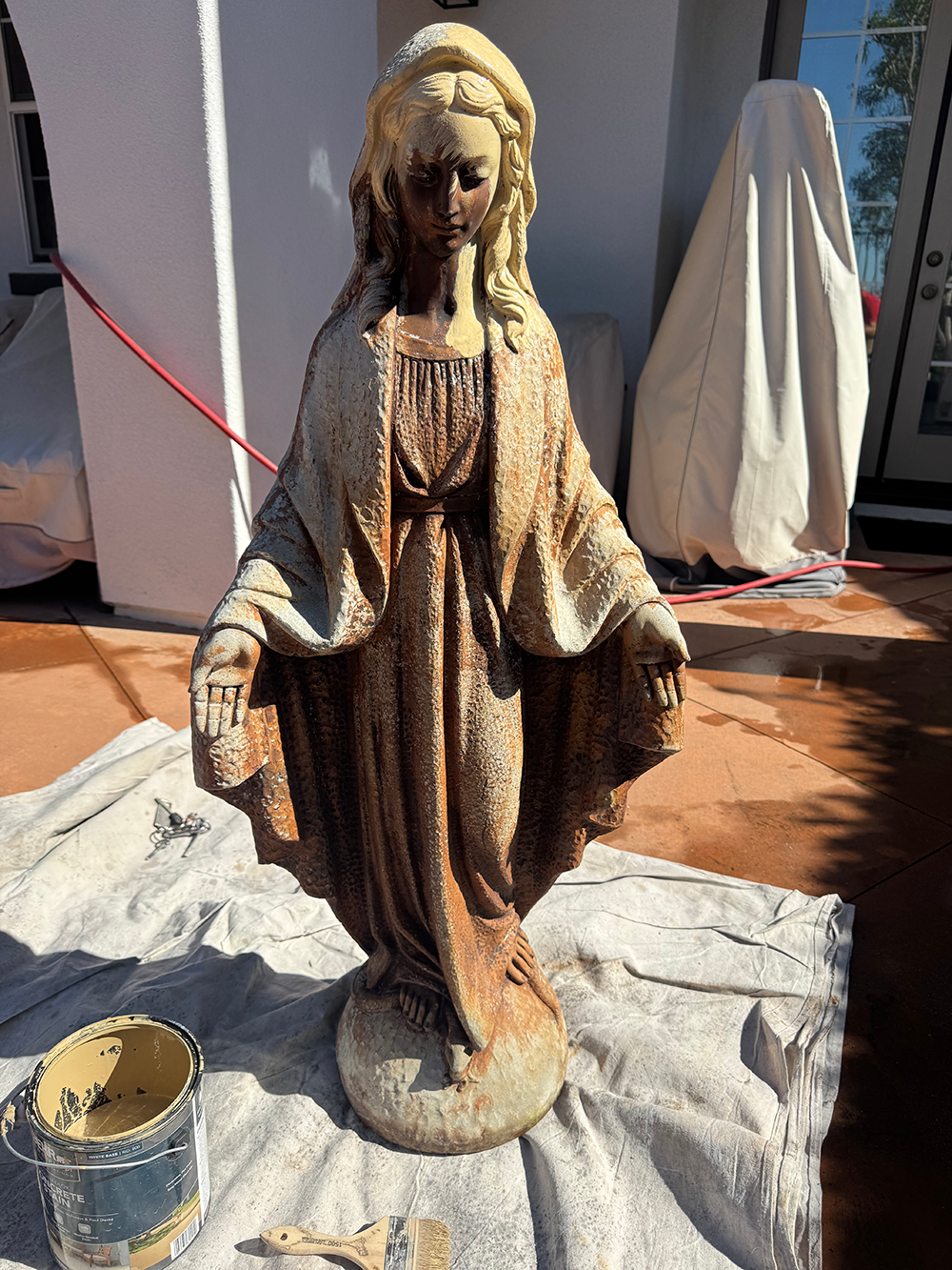 Statue of Virgin Mary, weathered, outdoors. Standing with open hands, rust-colored streaks.