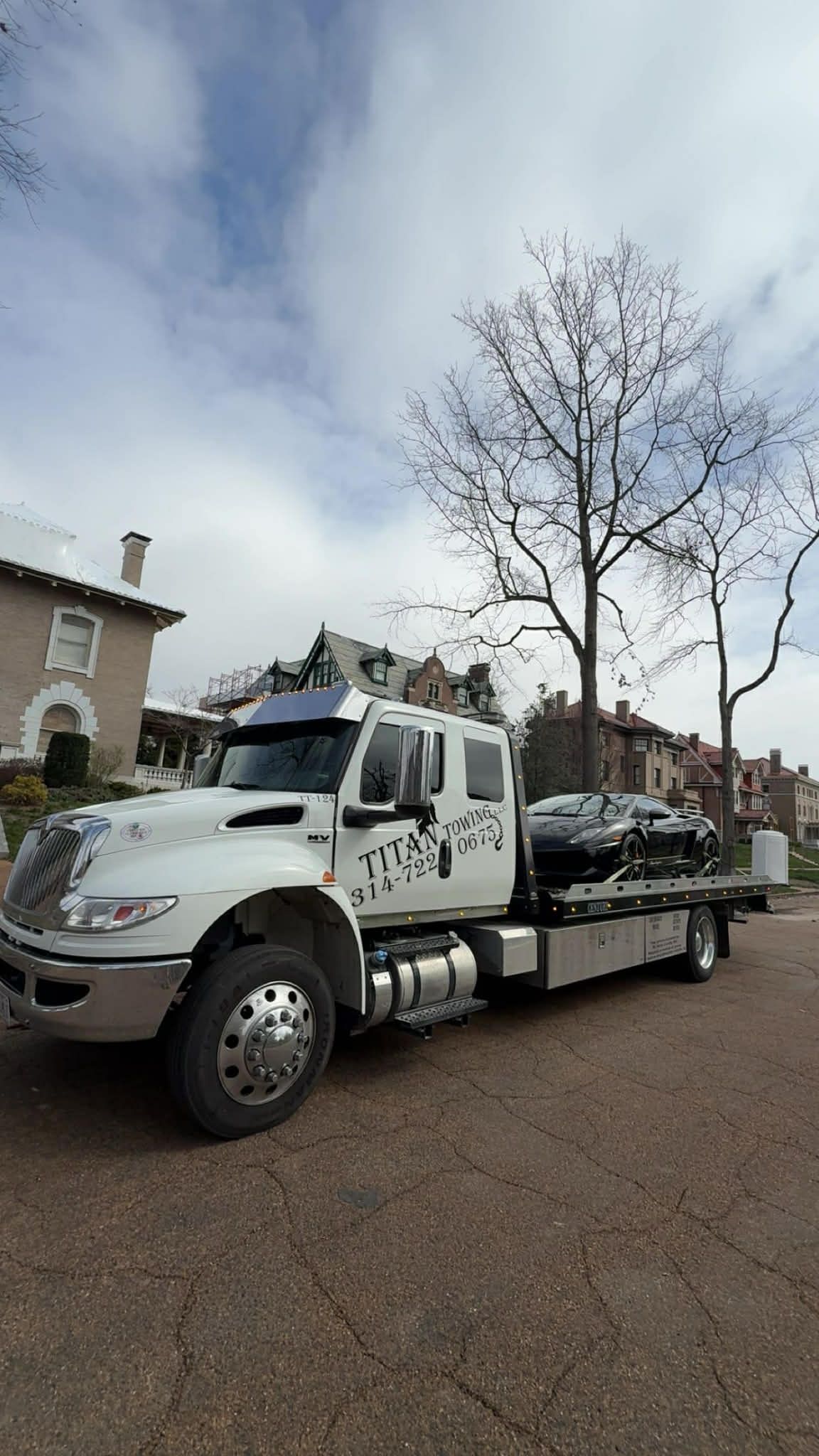 A white flatbed tow truck carrying a black car parked on a paved lot with a building and bare trees in the background.