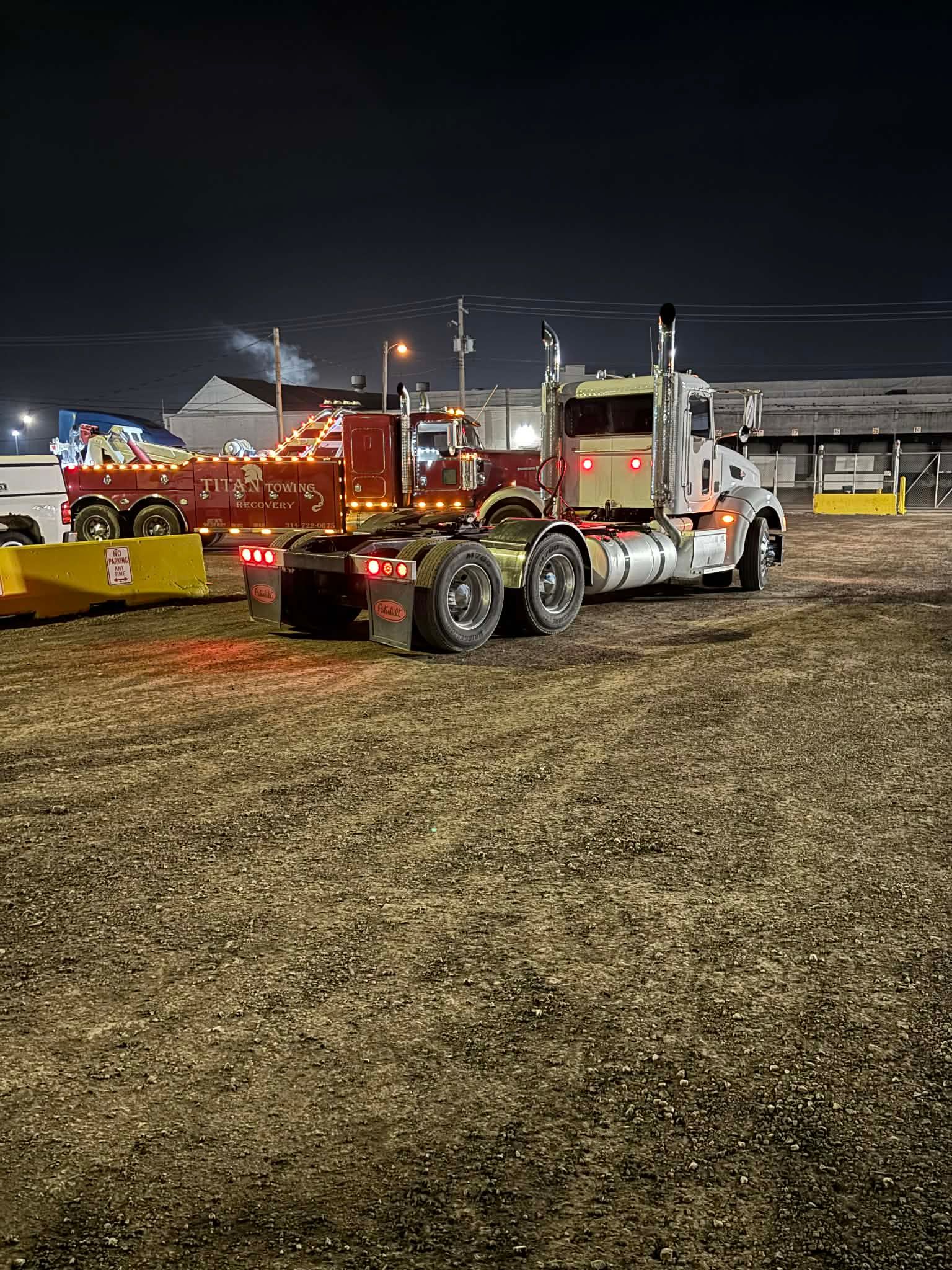 A white semi-truck and a red tow truck parked in a large, dark, gravel-covered lot at night.