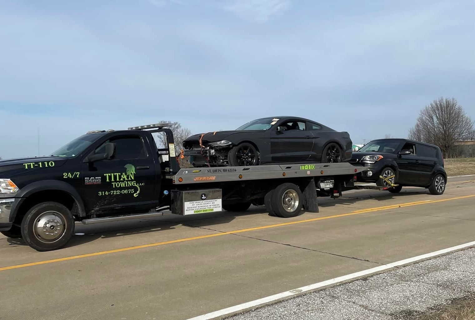A black tow truck hauling a damaged black sports car and a black SUV on a highway. The sky is blue.