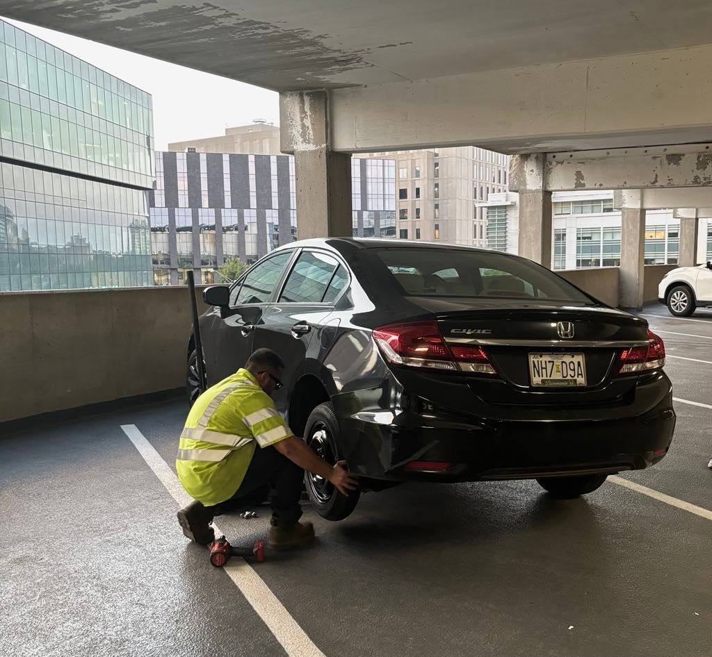 Man changing a tire on a black car in a parking garage; he wears a yellow safety vest.