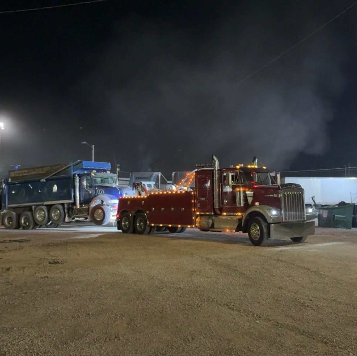 A maroon tow truck towing a blue dump truck at night.