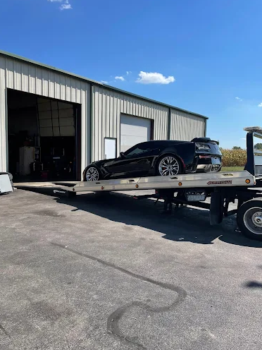 Black sports car on a flatbed tow truck outside a garage on a sunny day.