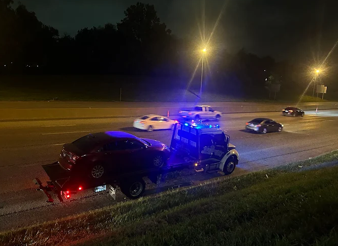 A tow truck with flashing blue lights is towing a damaged black car on a dark highway at night. Other cars are visible.