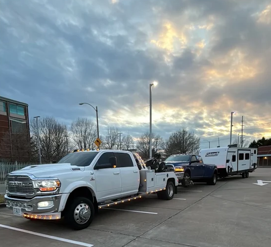 A white tow truck pulling a pickup truck and a camper trailer in a parking lot under a cloudy sky.