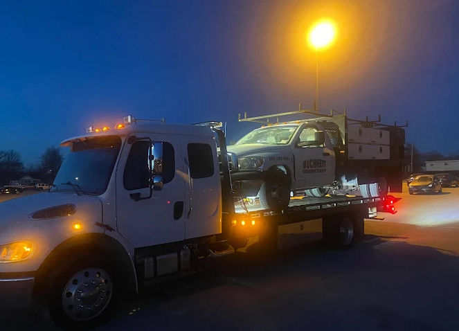White tow truck with a truck on its flatbed, illuminated by streetlights at dusk.