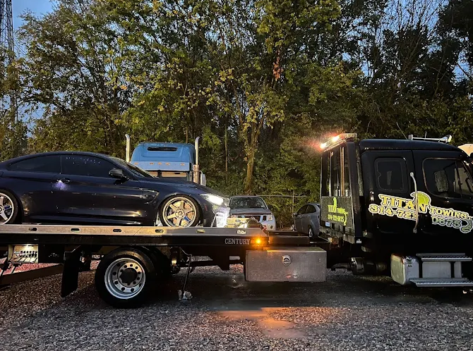 A dark car is being towed onto a flatbed truck at dusk. The tow truck is black with bright lights. Trees are in the background.