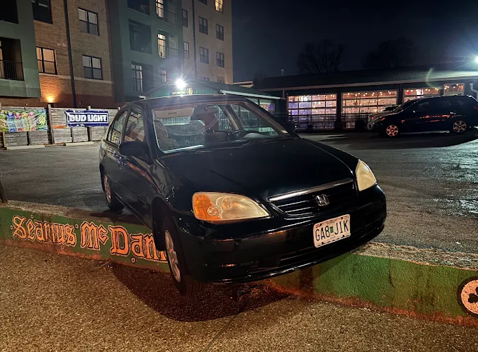 Black Toyota parked on a curb next to a green mural with lettering. Buildings and a lit bar are in the background.