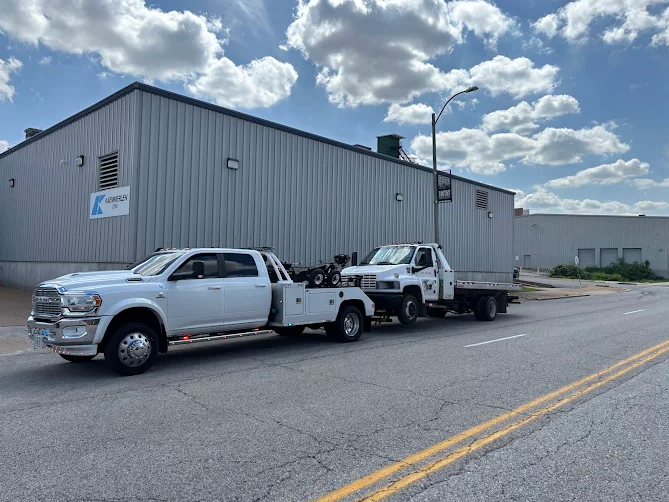 A white tow truck towing another flatbed truck on a city street, in front of a metal building under a partly cloudy sky.