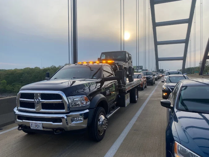 A black tow truck carrying a Jeep Wrangler on a bridge, with cars in traffic. The sun shines through the bridge's arches.