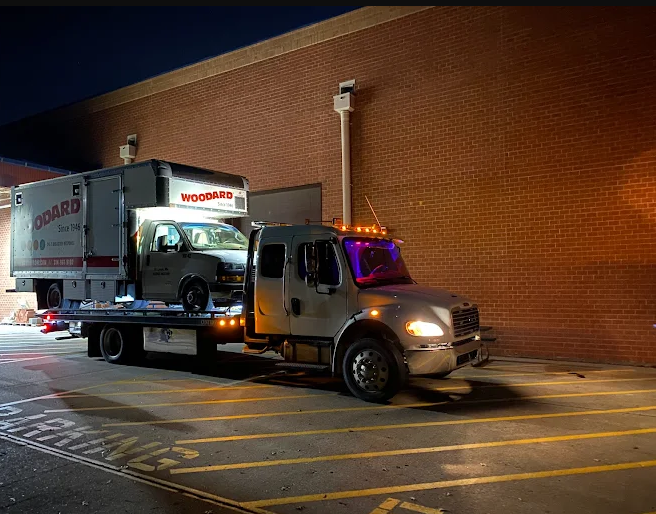 A tow truck at night with a car loaded on the flatbed, parked next to a brick building.