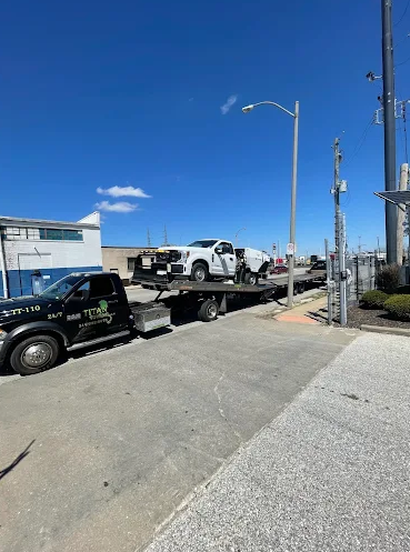 A white truck being transported on a flatbed trailer pulled by a black tow truck on a sunny day.