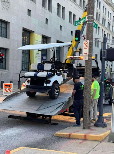 A white golf cart with a canopy is being loaded onto a tow truck in a city street. Two men stand nearby.