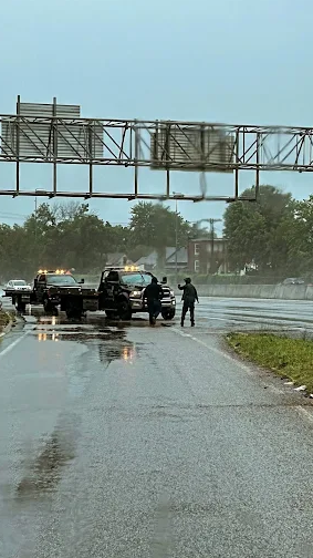 Two tow trucks and personnel on a rain-slicked highway. The trucks have flashing lights, and one person gestures.