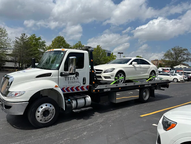 White tow truck carrying a white Mercedes-Benz on a sunny day in a parking lot.