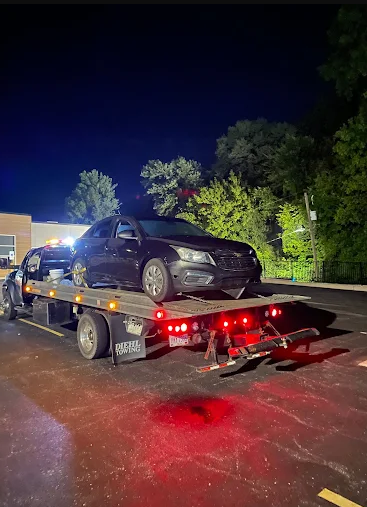 A black car is on a tow truck at night. The truck's lights are red, and it is parked in a lot with a dark sky and trees.