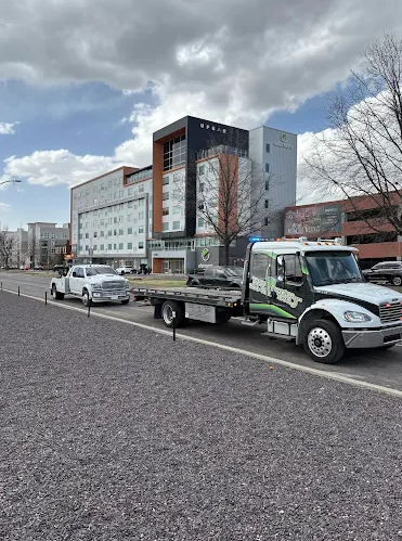 A flatbed tow truck towing a white classic car on a street in front of a modern multi-story building.