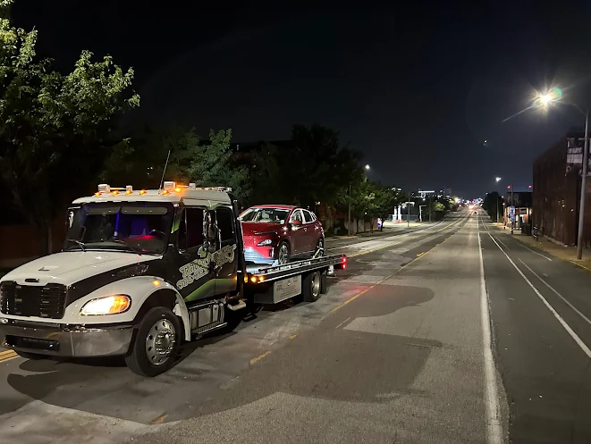 A tow truck with a red car on its flatbed is parked on a street at night, illuminated by streetlights.