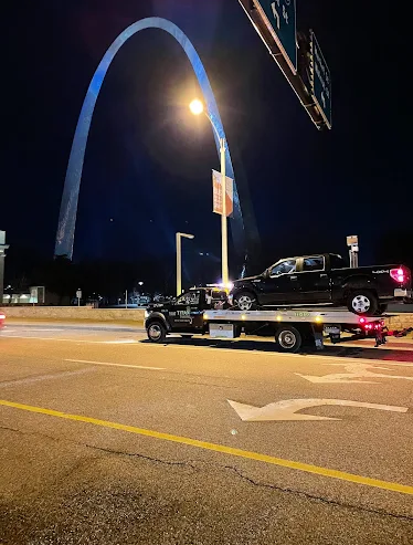 A black pickup truck is being towed at night in front of the Gateway Arch in St. Louis. The arch is lit up in blue.