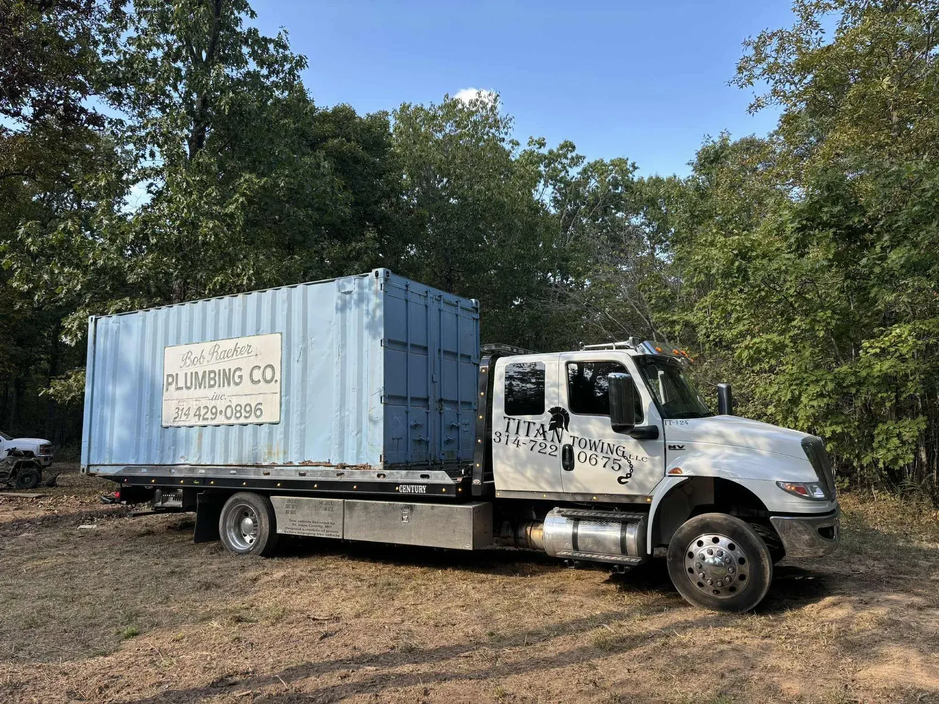 White plumbing truck with a blue shipping container on the flatbed, parked on dirt near trees.