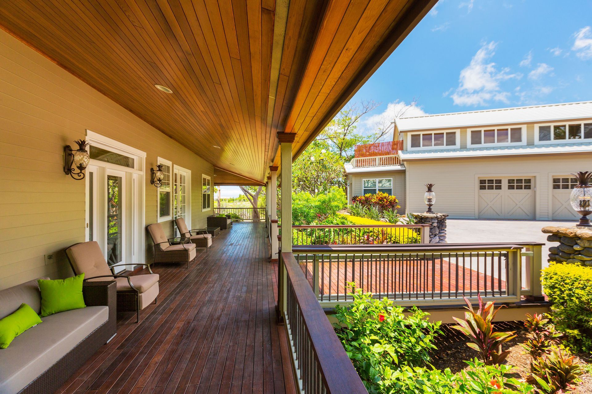 Covered wooden patio deck with seating, green cushions, and potted plants beside a bright house and garage.