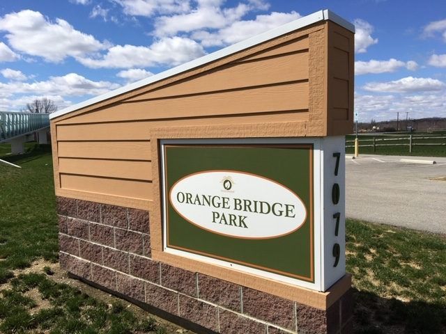 Sign for Orange Bridge Park with orange siding, green and white logo, and brick base.