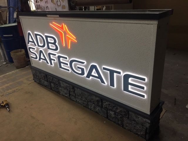 Lit-up sign for ADB SAFEGATE with orange logo and white-lit text, set in a stone and gray bordered display.