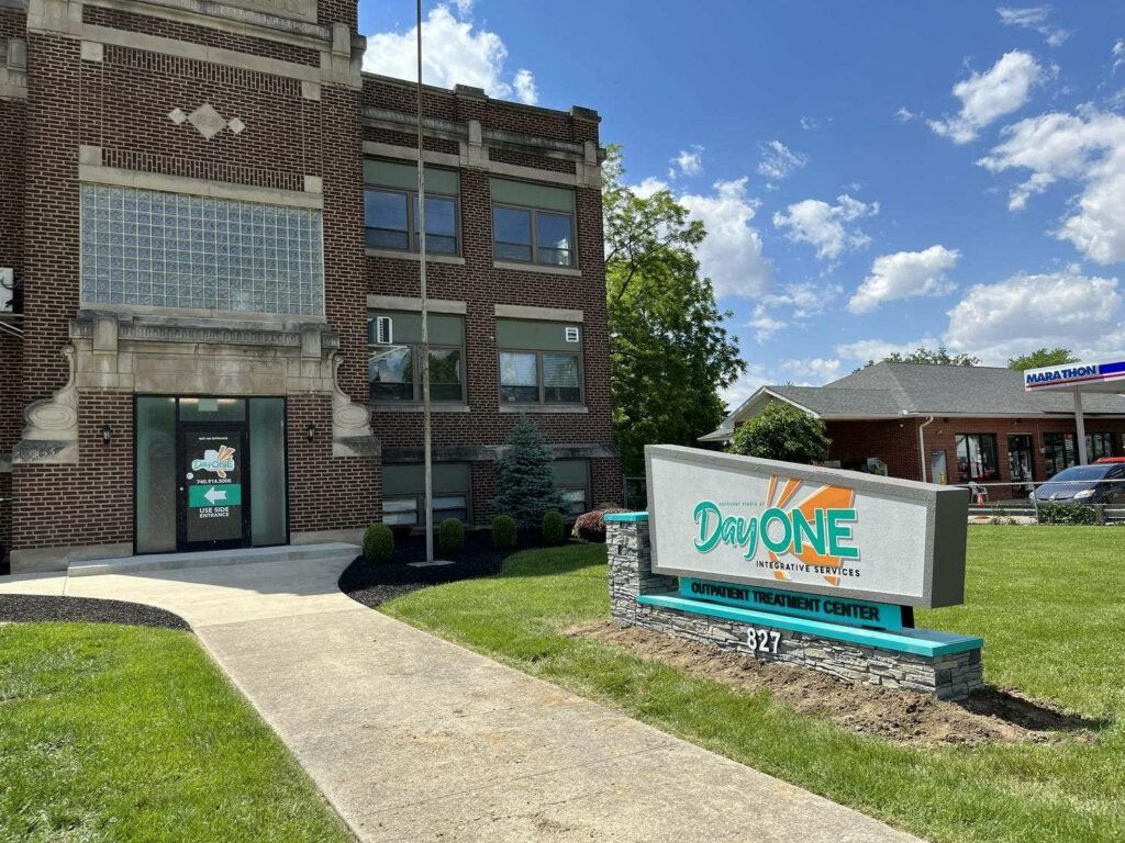 Day One addiction treatment center exterior. Brick building with sign, blue-green accents, and sidewalk.