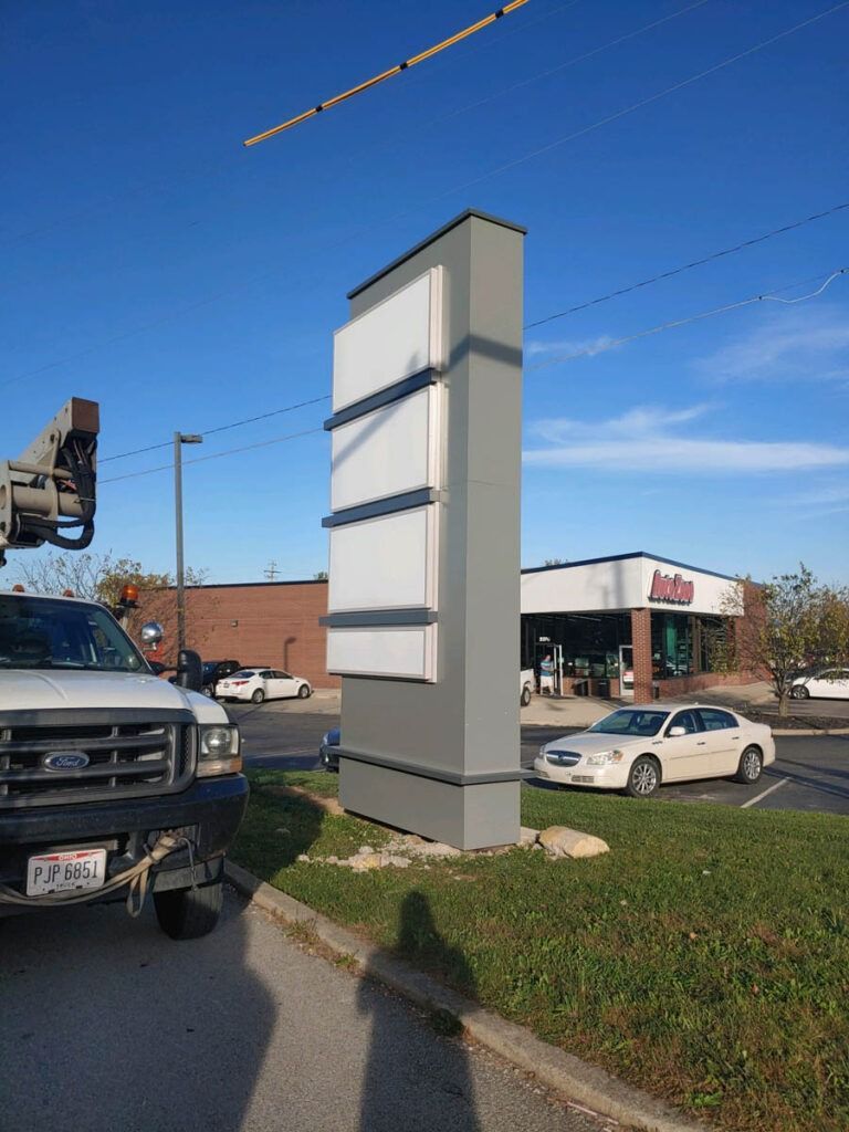Tall gray business sign with blank panels stands on green grass; truck and car in foreground.